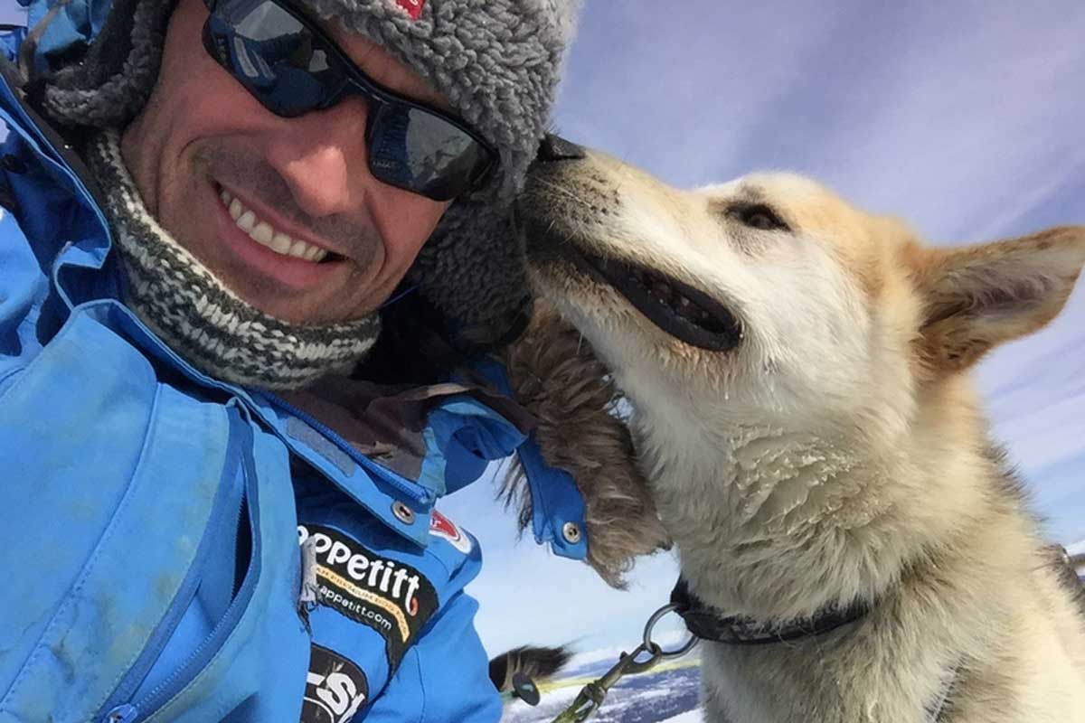 Smilende Appetitt ambassadør Thomas Wærner med lue og solbriller, blir snust på hodet av en av sine sledehunder under Finnmarksløpet.