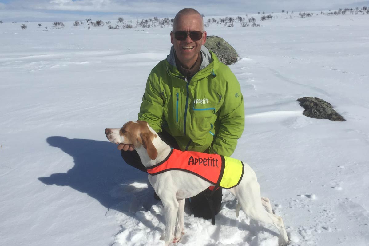 Appetitt ambassadør Rune Hoholm med grønn Appetitt-jakke smiler til kameraet, sittende i snøen sammen med sin fuglehund iført vest fra Appetitt.